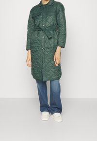 Abrigo verde acolchado largo con cintura ceñida, botones a presión y dos bolsillos en el pecho. Llevado sobre unos vaqueros acampanados azules y zapatillas blancas.