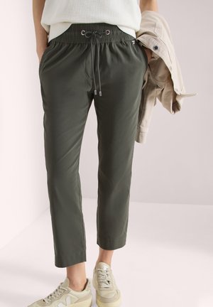 Person trägt dunkelgrüne Kordelhose und beigen Sneakers, hält eine beige Jacke in einer Hand.