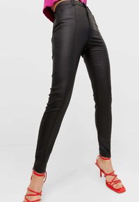 Pantalons skinny en simili cuir noir avec une taille haute, présentant un design ajusté et des détails zippés aux chevilles. Portés avec des talons rouges à lanières.