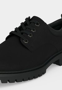 Chaussure noire en cuir à lacets avec œillets métalliques, semelle épaisse et robuste, talon bas, vue en gros plan à angle.