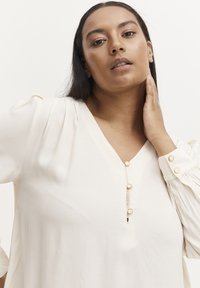 Blusa color crema con scollo a V e patta bottonata. Presenta spalle plissettate e maniche con polsini bottonati in tonalità oro. Tessuto morbido.