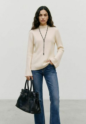 Femme portant un pull côtelé crème et un jean bleu tenant un grand sac à main en cuir noir avec deux poignées sur un fond uni.