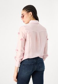 Light pink, long-sleeve shirt with small bow details on sleeves; paired with dark denim jeans. The fabric has a slightly textured appearance.