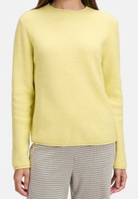 Femme portant un pull côtelé jaune doux et un pantalon à carreaux beige, photographiée des épaules aux hanches sur fond blanc.