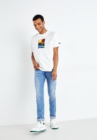 Camiseta blanca de algodón con un diseño gráfico colorido, combinada con jeans de color azul claro y zapatillas blancas con acentos verdes.