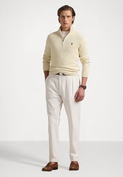 Pull à col zippé crème, pantalon blanc, mocassins bruns. Présente un design ajusté, des poignets côtelés et un petit logo sur la poitrine.