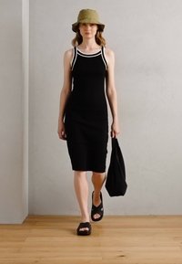 Robe noire ajustée avec des bordures blanches, sandales plates, chapeau seau vert olive et un sac fourre-tout noir. Texture lisse, design minimaliste. Sol en bois.