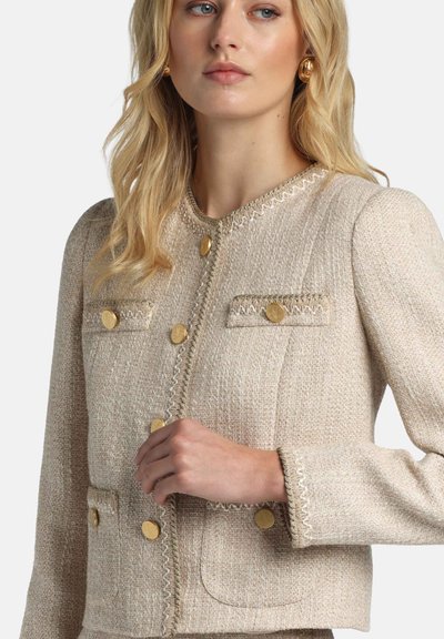 Beige tweed jacket with gold buttons, textured fabric, embellished pockets, and decorative stitching along the neckline and cuffs.