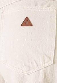 Beige denim pocket with a small brown triangular leather patch stitched near the top center.