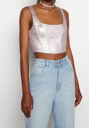 Kobieta w liliowym metalicznym bezrękawniku typu crop top, jasnoniebieskich jeansach z wysokim stanem i kwiatowym naszyjnikiem typu choker na białym tle.