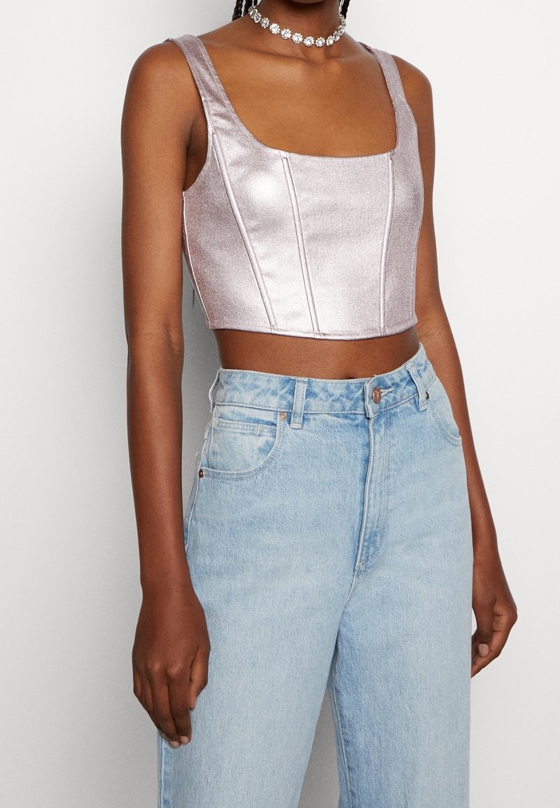 Femme portant un crop top métallisé lilas sans manches, un jean taille haute bleu clair et un collier ras de cou floral sur fond blanc.