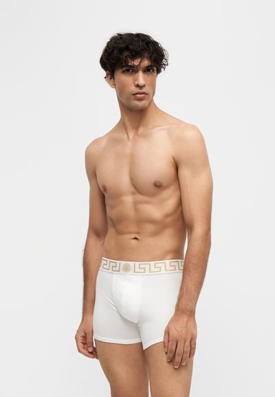 Jeune homme aux cheveux bouclés foncés portant un boxer blanc avec une ceinture géométrique dorée, debout devant un fond clair uni.