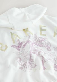 Valkoinen huppari, jossa on kultaisia ja vaaleanpunaisia grafiikoita, kukkakuvioita, avainyksityiskohtia ja brändin nimi lihavoidulla tekstillä. Pehmeä tekstuuri.