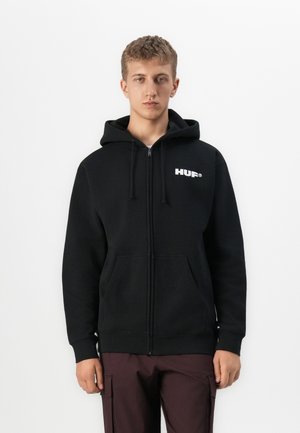 Young man wearing a black zip-up hoodie with "HUF" logo on chest and dark cargo pants, standing against a plain white background.