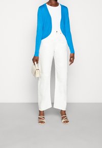 Blå långärmad cardigan över en vit t-shirt, matchad med vita vida byxor och beige remsandaler, håller en liten krämfärgad väska.