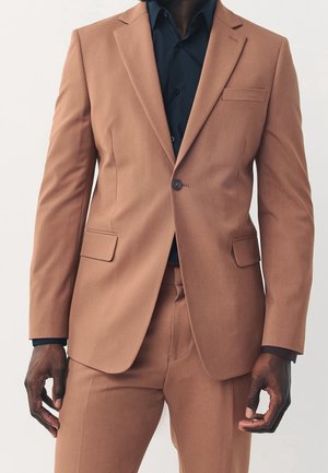 Homme portant un costume marron clair ajusté avec une veste à un bouton sur une chemise noire, debout devant un fond uni.