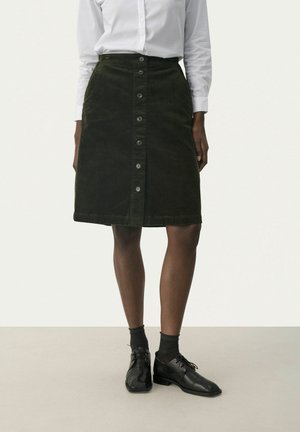 Vrouw met een donkergroene corduroy rok met knopen aan de voorkant, een wit shirt met lange mouwen, zwarte sokken en zwarte veterschoenen, staand op een neutrale vloer.