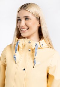 Chaqueta amarilla impermeable hecha de material suave, con capucha ajustable, detalles a rayas azules y blancas, y cierres a presión.