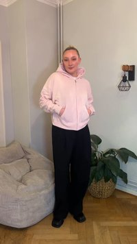 Camisola com capuz rosa claro com fecho de correr e bolsos frontais, combinada com calças pretas de perna larga e sapatos pretos. No fundo, incluem-se uma planta e uma poltrona texturada.