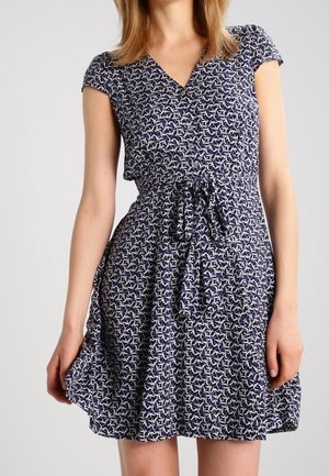 Frau trägt ein marineblaues Kleid mit kurzen Ärmeln, weißem Blumenmuster, durchgeknöpft vorne und gebundenem Taillengürtel.