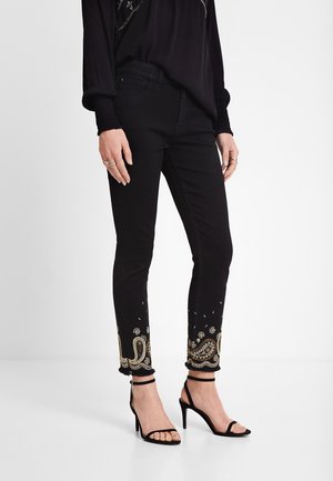 Svarta denimjeans med fransdetalj har intrikata beige och metallisk broderi vid fållarna. Matchas med svarta remsandaler.