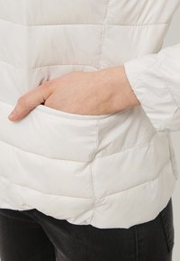 Hand inskjuten i sidofickan på en vit quiltad jacka som bärs över svarta byxor.