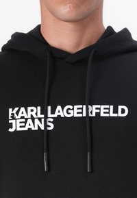 Čierna mikina s kapucňou featuring biely grafický potlač s nápisom "KARL LAGERFELD JEANS." Obsahuje čierne šnúrky s plastovými špičkami. Mäkká textúra tkaniny.