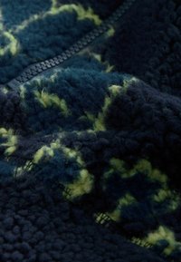 Texturovaná fleece tkanina v tmavě námořnické barvě s jasně zelenými akcenty. Obsahuje viditelný černý zip podél švu. Měkká a nadýchaná textura.