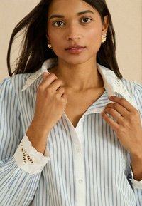 Vaaleansininen ja valkoinen raidallinen nappipaita, jossa valkoinen kaulus pitsikoristeilla ja liepeissä siksakreunukset.