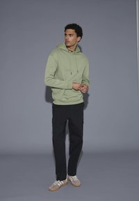 Lichtgroene hoodie met verstelbare trekkoorden, kangoeroezakken en geribde manchetten. Gepaard met zwarte cargobroeken en grijze sportschoenen.