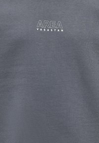 Sivkasta, teksturirana tkanina s besedami "AREA VASASTAN" v beli minimalistični pisavi na sredini. Preprost dizajn, krojaška ovratnik.