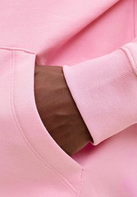 Sweat-shirt rose avec poignets côtelés et poche kangourou, montrant une main dans la poche. Texture lisse et détail de couture nette visibles.