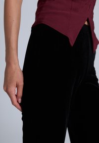 Černé sametové kalhoty v kombinaci s vínově červenou košilí. Košile má zakulacený lem a je přiléhavá, zatímco kalhoty jsou elegantní a hladké.
