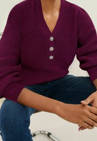 Suéter de punto morado con cuello en V, que presenta tres botones con forma de flor en la parte delantera. Puños acanalados y ajuste holgado.