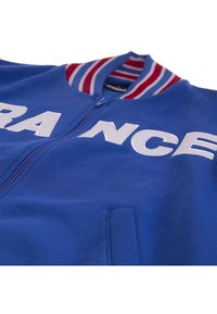 Blauwe ritsjack met witte letters "FRANKRIJK", geribbeld rood en blauwe kraag en zijzakken. Gemaakt van een zachte, gestructureerde stof.