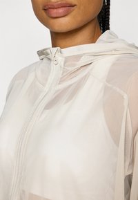 Lättviktig, genomskinlig zip-up jacka i off-white, med huva, slät yta och subtila sömdetaljer. Minimalistisk design.