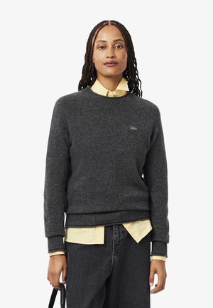 Grauer Wollpullover mit rundem Ausschnitt, gerippten Bündchen und Saum, über einem gelben Shirt getragen; verfügt über ein kleines Logo auf der Brust.