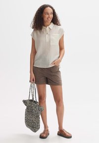 Helles beige Kurzarmhemd mit Kragen und einer Brusttasche, kombiniert mit braunen Shorts und einer gemusterten Tragetasche, getragen mit braunen Sandalen.