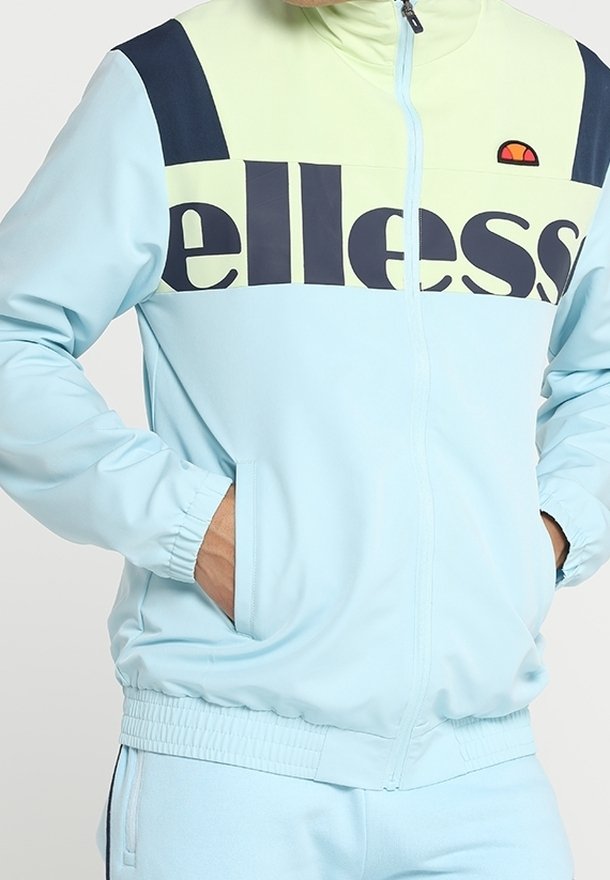 Veste zippée bleu clair avec un design en blocs de couleurs marine et vert clair. Caractéristiques d'un grand logo "ellesse", de poches zippées et de poignets élastiques.