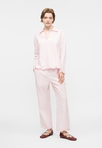 Νέα γυναίκα που φοράει ανοιχτό ροζ μακρυμάνικη μπλούζα και ταιριαστό παντελόνι με κυματιστά τελειώματα, στέκεται με τα χέρια χαλαρά, καφέ παπούτσια με κορδόνια.