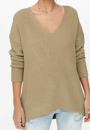 Pull beige en maille à col en V avec manches longues porté sur un jean bleu clair déchiré sur une personne debout, les bras détendus.