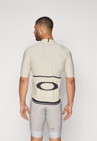 Maillot de cyclisme à manches courtes en beige clair avec un logo noir au dos et un short ajusté gris avec des accents noirs et bleu clair.