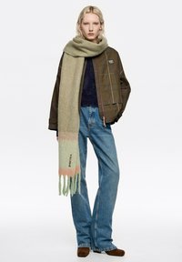 Jasnozielony szal o miękkiej fakturze, z gradientem kolorów i ozdobnymi frędzlami. Nosi się go z ciemną, wzorzystą kurtką i niebieskimi dżinsami.