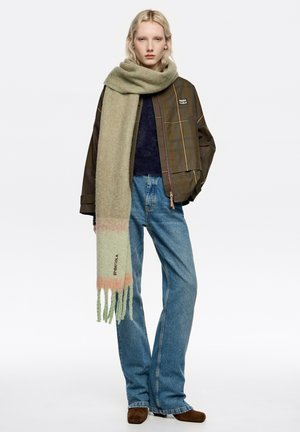 Jasnozielony szal o miękkiej fakturze, z gradientem kolorów i ozdobnymi frędzlami. Nosi się go z ciemną, wzorzystą kurtką i niebieskimi dżinsami.