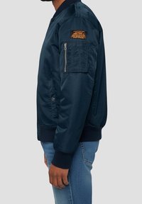 Homme portant une veste bomber bleu foncé avec une poche zippée et une étiquette sur la manche supérieure, associée à un jean bleu clair.