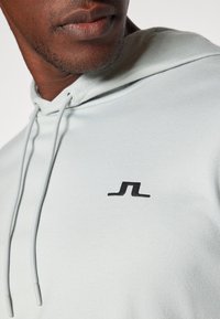 Moleton cinza-claro com um tecido texturizado, capuz com cordão e um pequeno logótipo preto no peito. Apresenta detalhes de costura subtis.