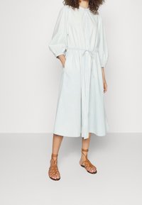 Vestito camicia azzurro chiaro con cintura in vita, maniche a palloncino e tasche laterali; abbinato a sandali marroni con cinturini. Tessuto morbido.