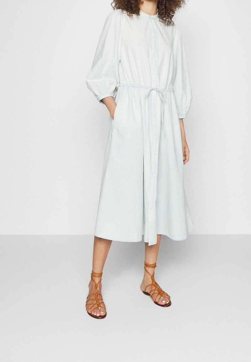 Vestito camicia azzurro chiaro con cintura in vita, maniche a palloncino e tasche laterali; abbinato a sandali marroni con cinturini. Tessuto morbido.