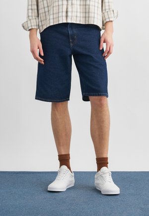 Uomo che indossa pantaloncini di denim blu scuro, sneakers bianche, calzini marroni e una camicia a quadri a maniche lunghe arrotolata, in piedi su un tappeto blu.