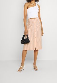 Top corto bianco con smock abbinato a una gonna a righe pesca e bianche lunga fino al ginocchio, con bottoni frontali. La modella tiene una borsa intrecciata nera.